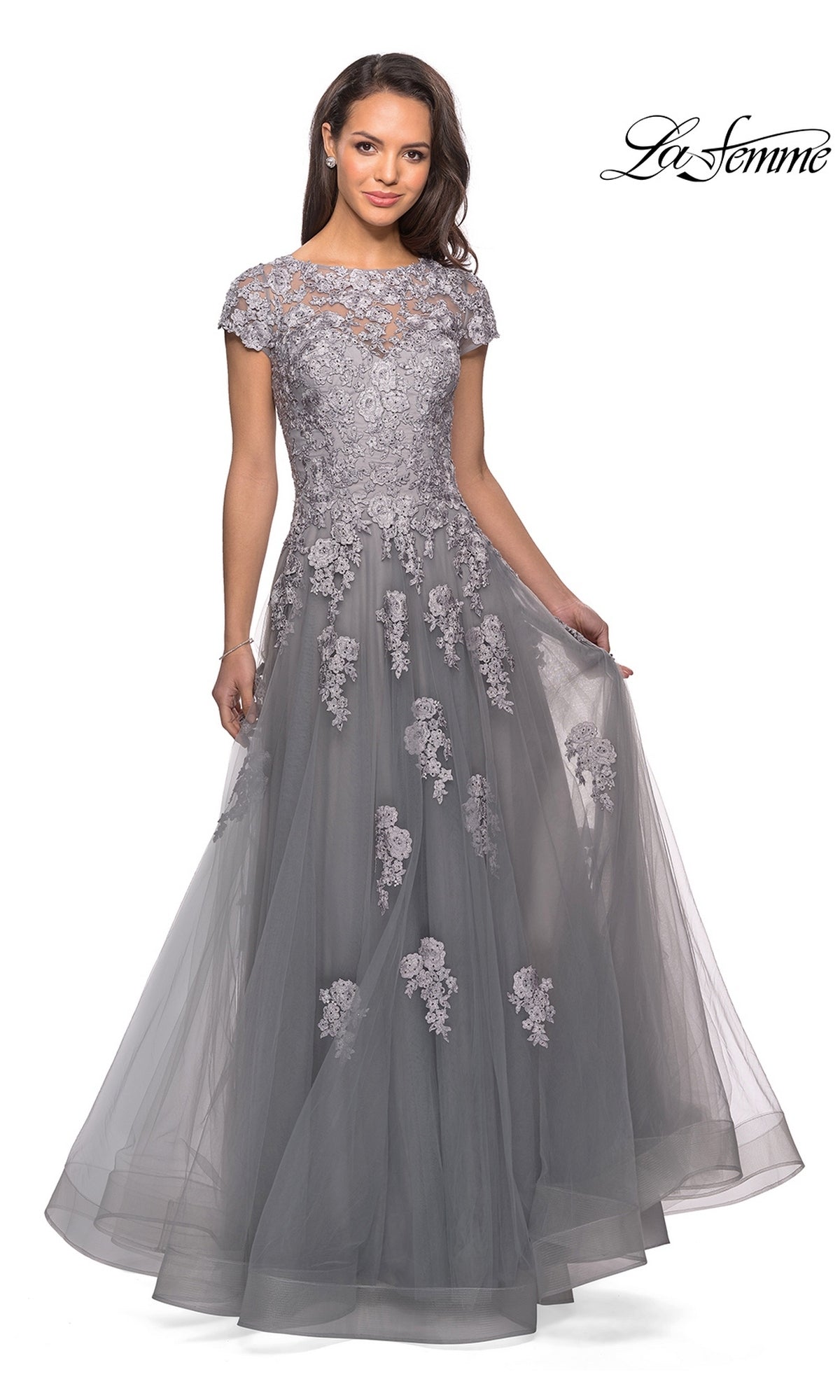 A woman models the Long Formal Dress: La Femme 26907—a floor-length, short-sleeved grey lace gown adorned with embellished lace appliques and floral embroidery on an elegant A-line silhouette, featuring a flowing tulle skirt—posing against a white background with the “La Femme” logo in the top right corner.